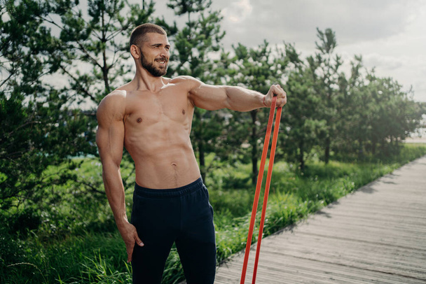 Strength and motivation concept. Muscular man with naked torso, has exercises with resistance band, performs training for biceps, looks into distance, poses outdoor. Sportsman uses expander. - Φωτογραφία, εικόνα
