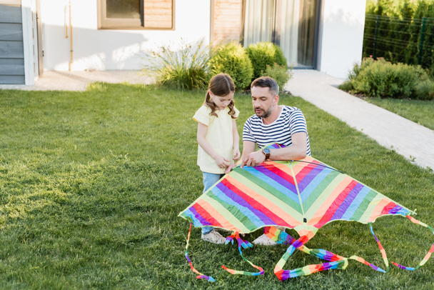 Full length of daughter standing near father assembling kite on lawn with house on background - Photo, Image