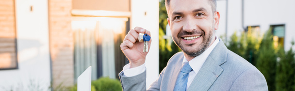 Portrait of happy real estate agent showing keys while looking at camera on blurred background, banner - Photo, Image