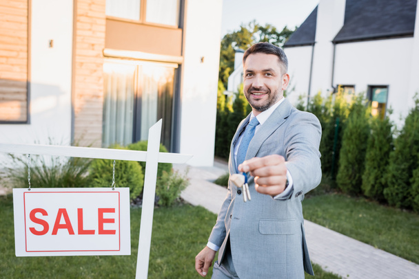 Happy broker looking at camera and standing near sign with sale lettering with blurred keys on foreground - Photo, Image