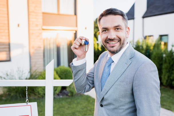 Happy broker showing keys while looking at camera on blurred background - Zdjęcie, obraz