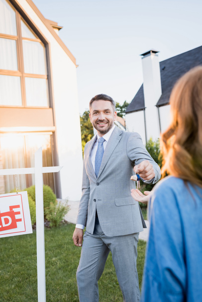 Smiling broker looking at camera while giving keys to blurred woman on foreground - Photo, Image