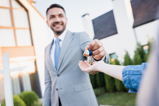 Smiling real estate agent giving keys to woman with blurred houses on background - Photo, Image