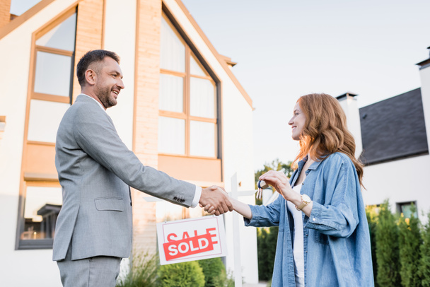 Side view of happy broker and woman with keys shaking hands near sign with sold lettering on blurred background - Foto, immagini