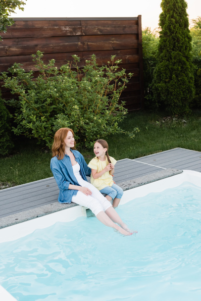 High angle view of happy mother and daughter sitting with legs in swimming pool near lawn with bushes on backyard - Foto, imagen