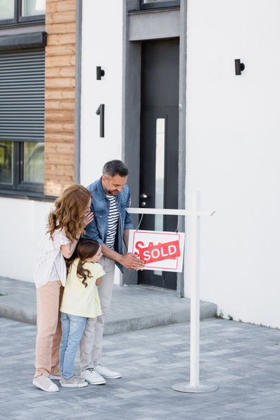 Full length of family hugging while looking at sign with sold lettering near house - Photo, Image