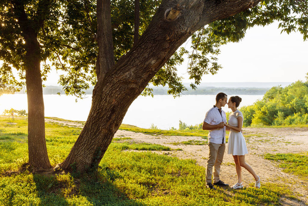 A man and a woman in white clothes stand under a huge old tree against the backdrop of the sunset. The noses of a woman and a man touch - Photo, Image