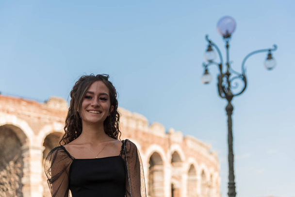 Полудлинный портрет молодой девушки с вьющимися каштановыми волосами, улыбающейся на улице. На заднем плане города, арена Верона, Италия. - Фото, изображение