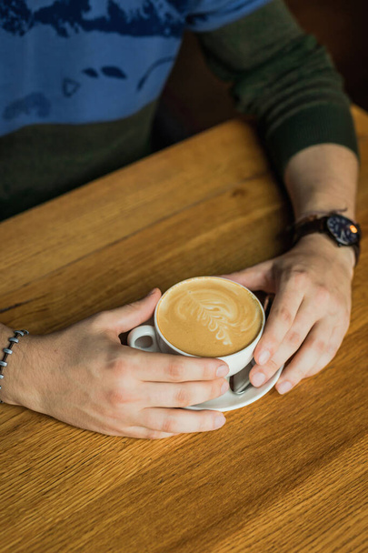 Male's hands holding a cup of cappuccino coffee, wooden table - Valokuva, kuva