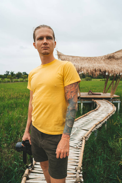 Man photographer in yellow t-shirt is walking at bamboo bridge in rice field - Photo, Image