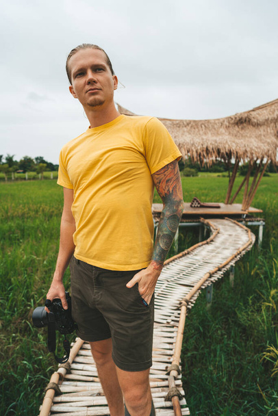Man photographer in yellow t-shirt is walking at bamboo bridge in rice field - Foto, Imagen