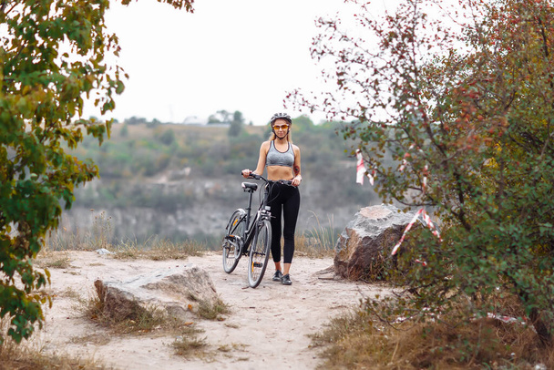 Fit young woman wearing sportswear standing with her bicycle on rocky background at autumn day - Foto, afbeelding