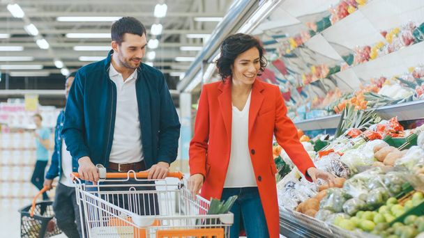 At the Supermarket: Happy Young Couple Chooses Organic Vegetables in the Fresh Produce Section of the Store. Boyfriend Pushes Shopping Cart while Girlfriend Picks up Groceries. - Φωτογραφία, εικόνα