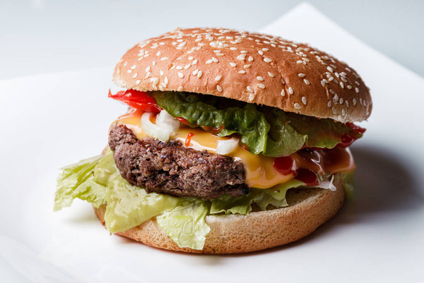 klasszikus burger marhahússal, sajttal, hagymával, ketchuppal, paradicsomszeletekkel és salátalevéllel, szezámmaggal és fehér tányéron - Fotó, kép