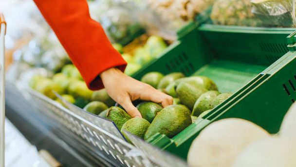 At the Supermarket: Close-up Shot of the Womans Hand Taking Avocado from the Fresh Produce Section and Places it into Shopping Cart. - Foto, Imagen
