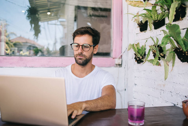 Serious bearded young male freelancer in casual clothes and eyeglasses working on laptop while sitting at table with glass of fresh beverage in modern street cafe - Fotografie, Obrázek
