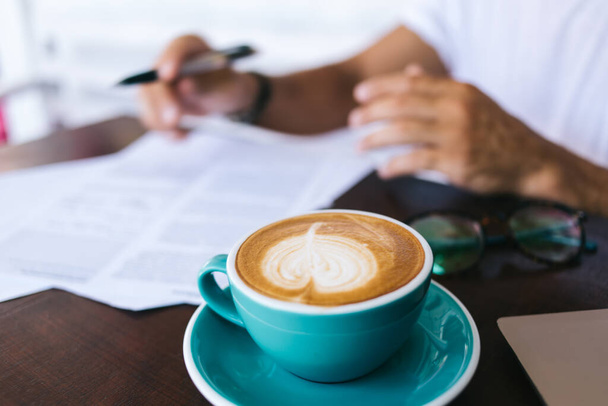 Cropped image of man writing on paper filling in application at working place with aroma hot caffeine beverage, selective focus on coffee mug with cappuccino standing on blurred background - Photo, image