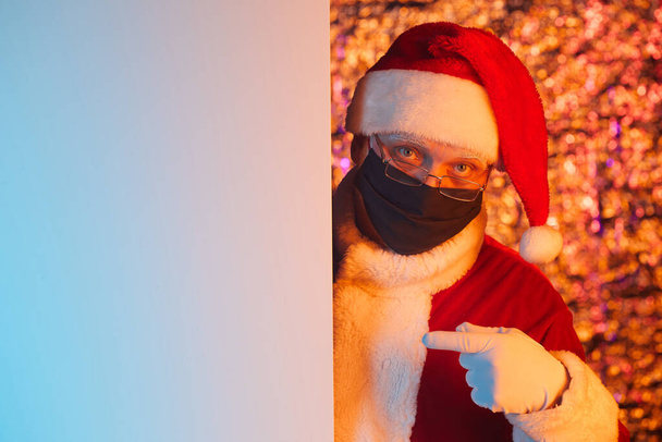 Portrait du Père Noël en masque de protection pointant vers une affiche vide recommandant quelque chose - Photo, image