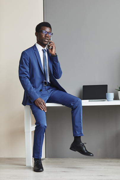 Portrait of African young businessman in blue suit and in eyeglasses sitting on the table and talking on mobile phone at office - 写真・画像