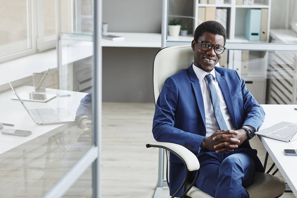 Portrét úspěšného afrického podnikatele, který se usmívá do kamery, zatímco sedí na židli v kanceláři - Fotografie, Obrázek