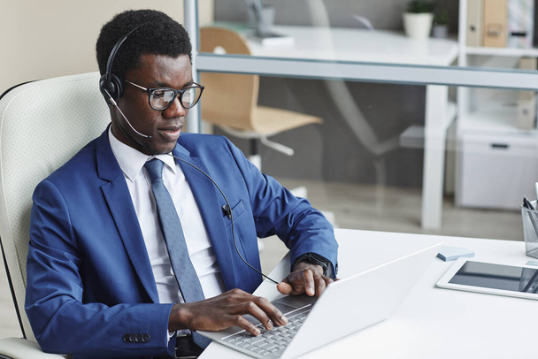 Африканский бизнесмен в наушниках печатает на клавиатуре компьютера на рабочем месте в офисе - Фото, изображение