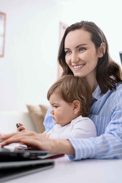 καυκάσια γυναίκα με παιδί κάθεται στο φορητό υπολογιστή, online ψώνια, εργασία από το σπίτι με παιδιά, κάθετη - Φωτογραφία, εικόνα
