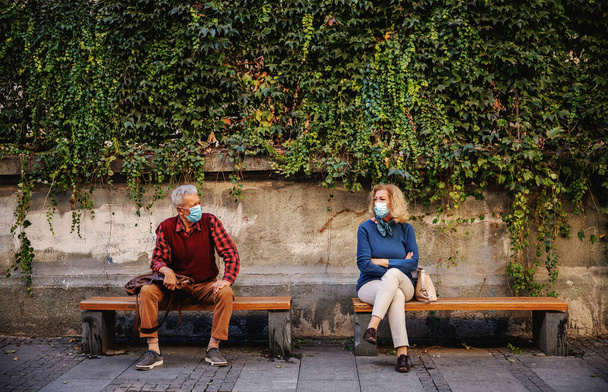 Personas mayores con máscaras protectoras sentadas en bancos y descansando. Las personas mayores estiman la distancia social. - Foto, imagen