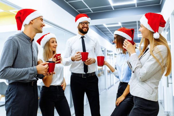 working team in Santa Claus hats, during a break in the office, greets the New Year and drinks a drink from red mugs - Foto, Imagen