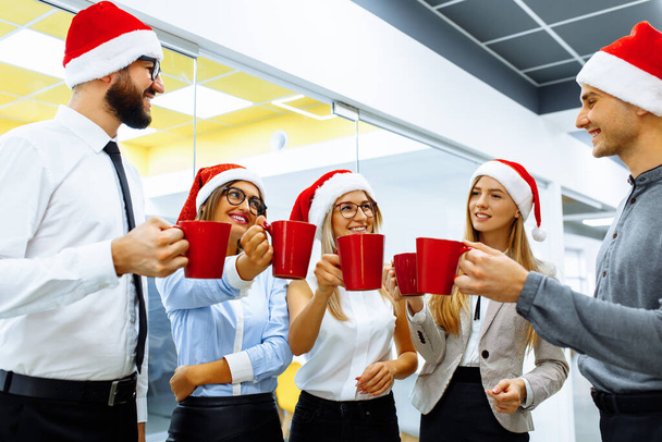 Група ділових людей у капелюхах Санта Клауса, Колеги святкують Новий рік в офісі, розважаються і п'ють напої з червоних кухлів. - Фото, зображення