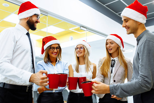 group of business people in Santa Claus hats, Colleagues celebrate New Year in the office, have fun and drink drinks from red mugs - Zdjęcie, obraz