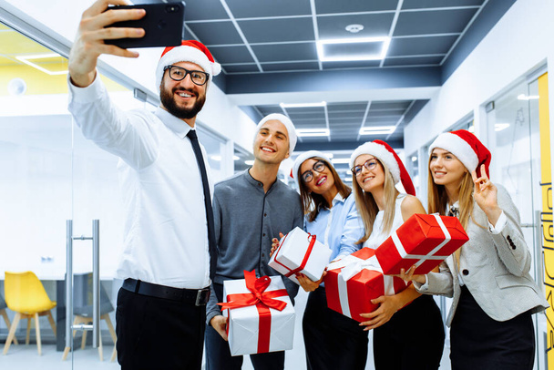 Happy group of businessmen in Santa Claus hats, with Christmas gifts are having fun celebrating New Year and taking selfie on a mobile phone - Fotografie, Obrázek
