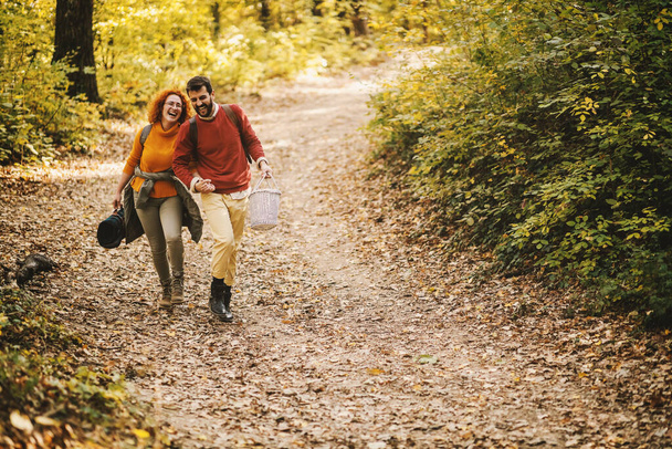 Ein junges fröhlich lächelndes verliebtes Paar hält sich an den Händen und geht an einem schönen Herbsttag in der Natur spazieren. Paar hält Picknickutensilien in der Hand. - Foto, Bild