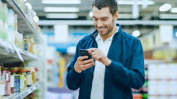 At the Supermarket: Handsome Man Uses Smartphone, Smiles while Standing at the Canned Goods Section. - Fotografie, Obrázek