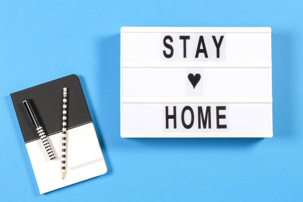 Caja de luz blanca con texto STAY HOME y cuaderno sobre fondo azul. Coronavirus, Covid-19, aprendizaje en línea, trabajo desde el hogar, cuarentena, concepto de distanciamiento social - Foto, imagen