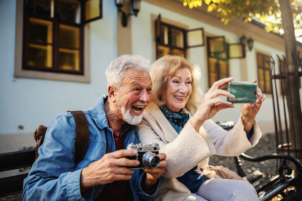 Bella coppia anziana seduta sulla panchina a farsi un selfie. L'uomo tiene la fotocamera mentre la donna tiene un telefono cellulare. - Foto, immagini