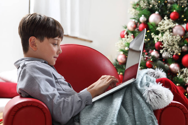 Veselé Vánoce, dítě doma s počítačem v blízkosti osvětleného a zdobeného stromu - Fotografie, Obrázek