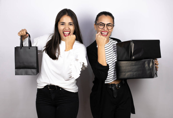 dwie młode piękne kobiety trzymające czarne torby na zakupy i pudełka podekscytowane wykonaniem gestu zwycięzcy z podniesionymi ramionami, uśmiechnięte i krzyczące o sukces. Czarny piątek - Zdjęcie, obraz