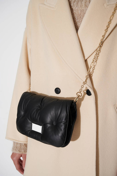 Κορίτσι με μπεζ παλτό και μαύρη δερμάτινη τσάντα σε χρυσή αλυσίδα. Πολυτελές υπόβαθρο μόδας. Κάθετη φωτογραφία. - Φωτογραφία, εικόνα