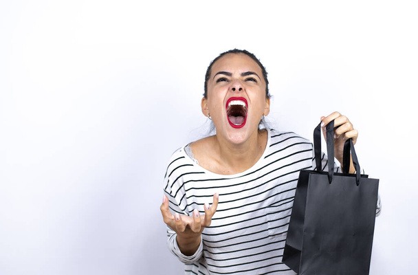 Νεαρή όμορφη γυναίκα κρατώντας μια μαύρη τσάντα ψώνια τρελός και τρελός φωνάζοντας και φωνάζοντας με επιθετική έκφραση και τα χέρια ψηλά. Έννοια απογοήτευση.. Μαύρη Παρασκευή - Φωτογραφία, εικόνα
