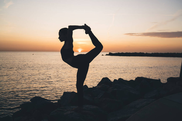 Full body side view of silhouette young female practicing yoga exercises at seaside on rocky shore of sea standing in Lord of the Dance pose at sunset - Photo, Image