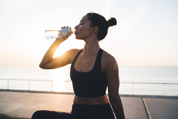 Caucasian female athlete in comfort tracksuit have workout break on embankment for drinking cold water and refreshness after aerobic pilates exercising, slim woman with perfect figure sitting at mat - Foto, afbeelding