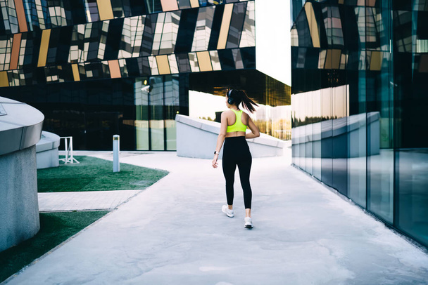 Back view of slim female athlete in casual sportswear and sneakers walking along sidewalk near modern buildings during training in headphones - Foto, Imagen