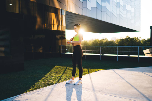 Fit female athlete in activewear standing on city street near glass building and listening to music in headphones while checking fitness tracker - Foto, Imagen