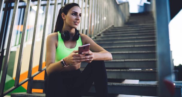 Low angle of ethnic thoughtful athletic female with cellphone in sporty outfit and headphones sitting on stairs and looking away while thinking over read message on summer day - Фото, зображення
