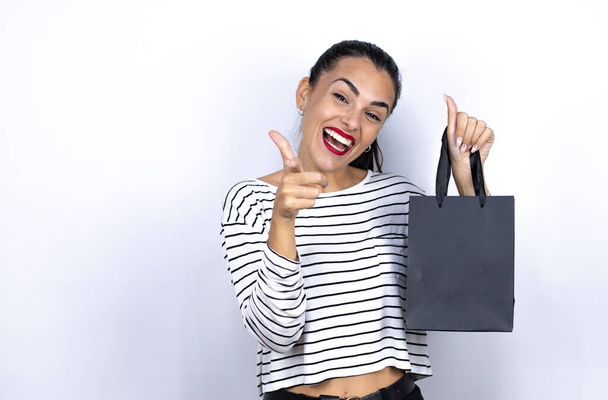 Joven mujer hermosa sosteniendo una bolsa de compras negro que apunta a usted y la cámara con los dedos, sonriendo positiva y alegre. Viernes Negro - Foto, imagen