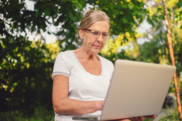 Technologia, koncepcja ludzi w podeszłym wieku - starsza staruszka pracująca online z laptopem na zewnątrz w ogrodzie. Praca zdalna, kształcenie na odległość.  - Zdjęcie, obraz