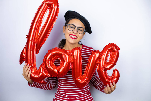Mladá krásná brunetka žena nosí francouzský baret a brýle přes bílé pozadí drží slovo láska balón - Fotografie, Obrázek