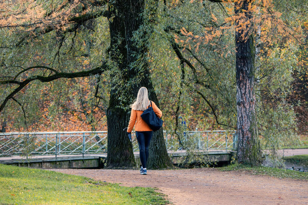オレンジのジャケットを着た美しいブロンドが秋の路地を歩いている。後ろからの眺め. - 写真・画像