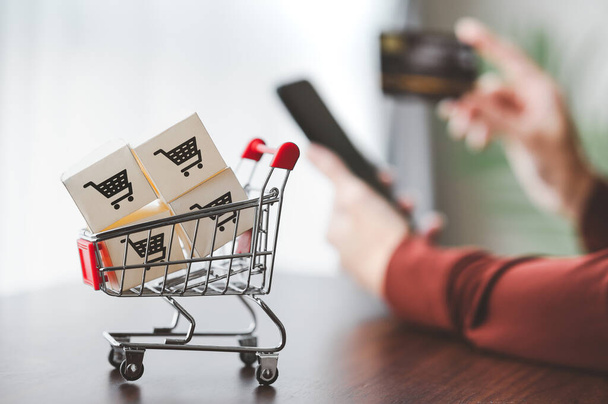 Papírdobozok bevásárlókosár logóval egy bevásárlókocsiban kézben tartott okostelefonnal és hitelkártyával történő kézbesítésre bacground-ban az online vásárláshoz - Fotó, kép