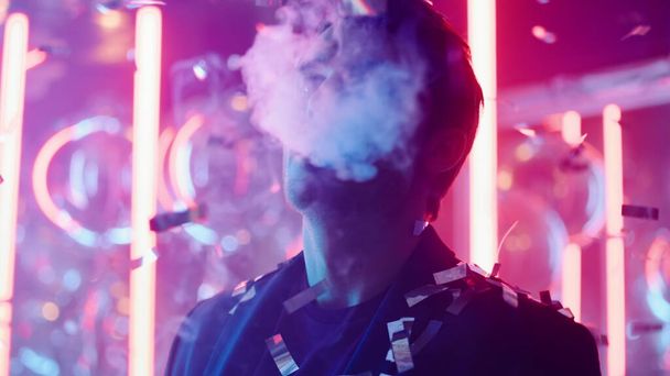 Young man smoking under confetti at party. Guy blowing smoke in nightclub - Foto, Bild